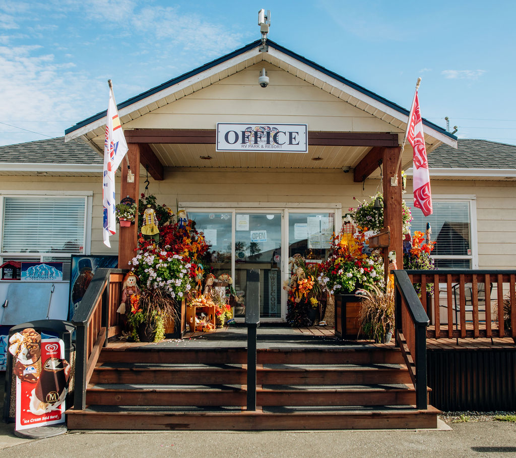 A small building with a sign reading "Office" above the entrance, decorated with autumn-themed flowers, plants, and scarecrows. Flags are on each side, and an ice cream freezer stands by the stairs.