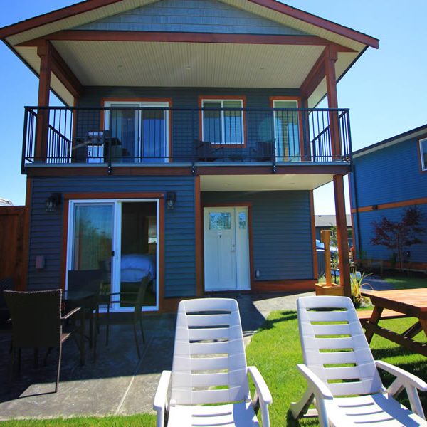 A two-story house with a covered balcony and patio, outdoor dining set, picnic table, and two white lounge chairs on a green lawn under a clear blue sky.