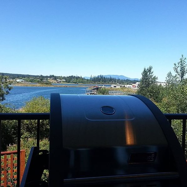 A shiny barbecue grill on a balcony overlooks a river, trees, and distant mountains under a clear blue sky.