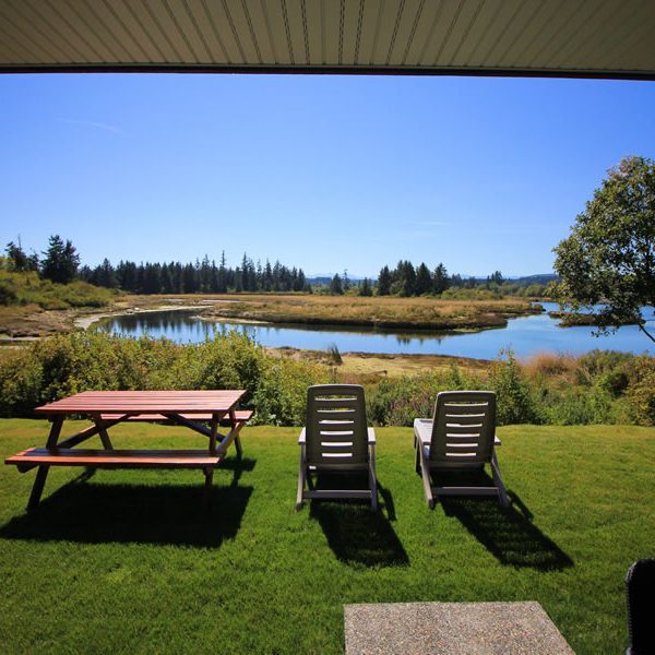 A grassy backyard with two lounge chairs and a picnic table overlooks a calm river or lake, surrounded by trees and clear blue skies on a sunny day.