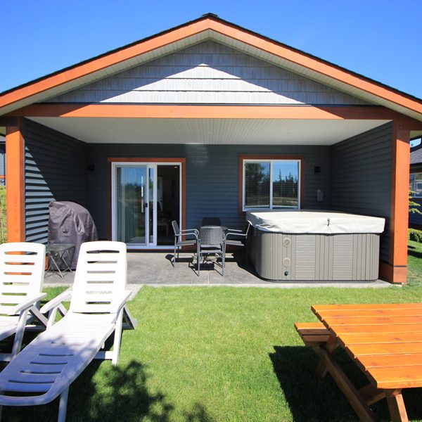 A modern backyard with a covered patio, outdoor furniture, a barbecue grill, and a hot tub. In the foreground are white lounge chairs on grass, next to a wooden picnic table, under a clear blue sky.